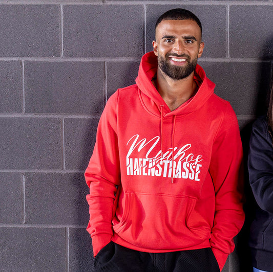 Ramien Safi trägt roten Rot-Weiss Essen Hoodie mit Kangarootasche, Ton-in-Ton Kordeln und Kordelenden, sowie rotes Gummi-Patch hinten links; mit weißem Mythos Hafenstrasse Druck zentriert auf Brust