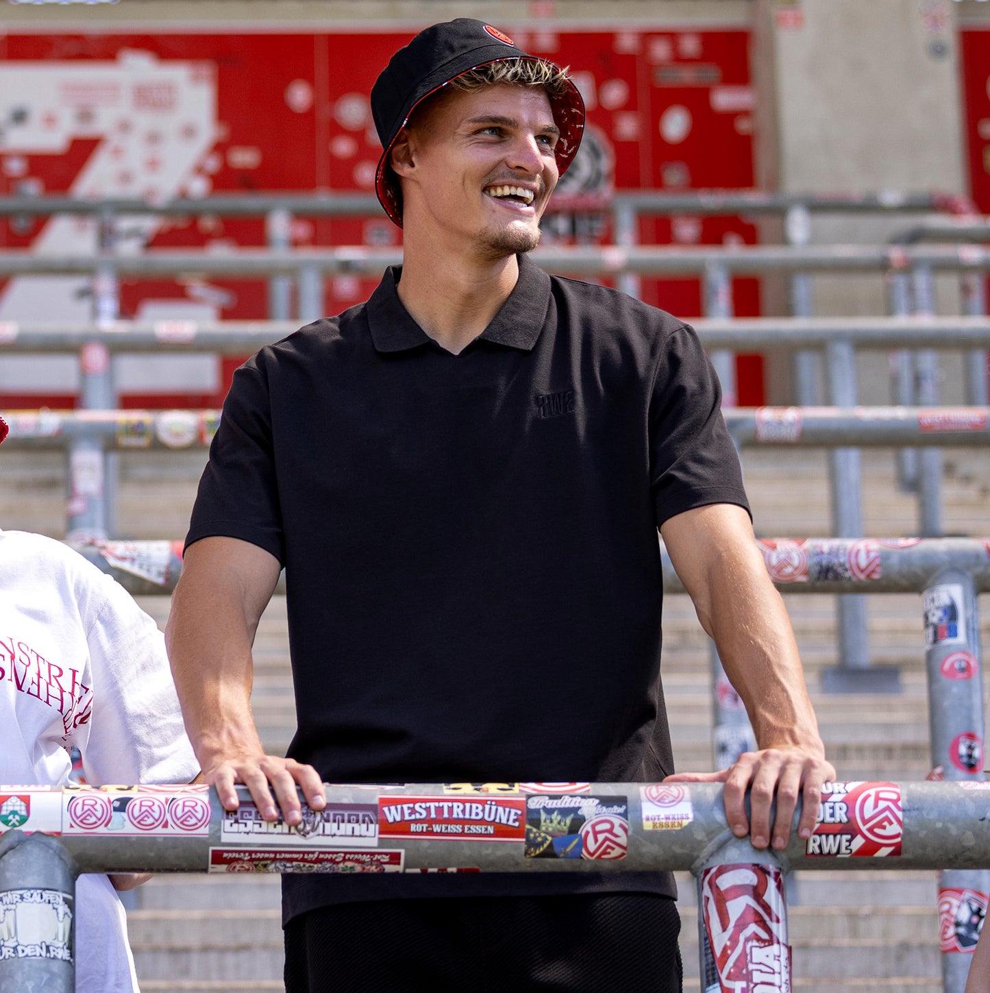Marek Janssen trägt schwarzes oversize Polo mit V-Ausschnitt und "RWE" Stickerei in schwarz auf linker Brust, keine Knopfleiste