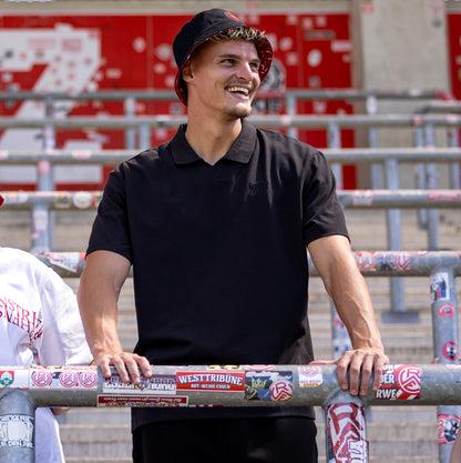Marek Janssen trägt schwarzes oversize Polo mit V-Ausschnitt und "RWE" Stickerei in schwarz auf linker Brust, keine Knopfleiste
