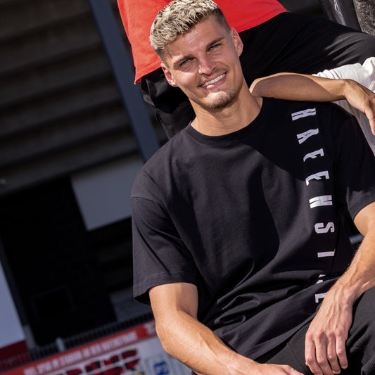 Marek Janssen trägt schwarzes Oversize T-Shirt mit vertikalem, großen weißen "HAFENSTRASSE" Druck auf Herzseite
