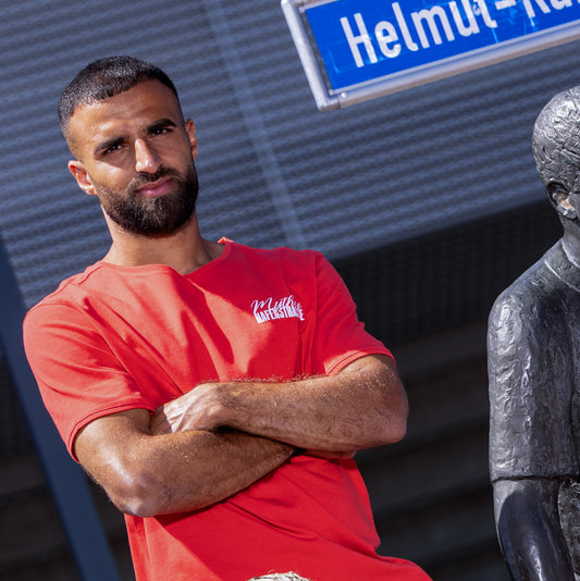 Ramien Safi trägt rotes T-Shirt im Regular Fit mit einem weißen "Mythos Hafenstrasse" -Druck auf der linken Brust. Der Druck ist eine Mischung aus Kalligraphie-Schrift (Mythos) und Rot-Weiss Essen-Schrifttyp (blockig; Hafenstrasse). Mit Rippbündchen am Ausschnitt und Ärmeln.