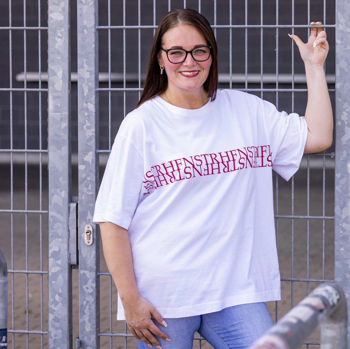 Jasmin trägt weißes Oversize Rundhals T-Shirt mit weißer Rippkante am Ausschnitt, rotes Nackenband, von Armloch zu Armloch roter Druck "HFNSTR"