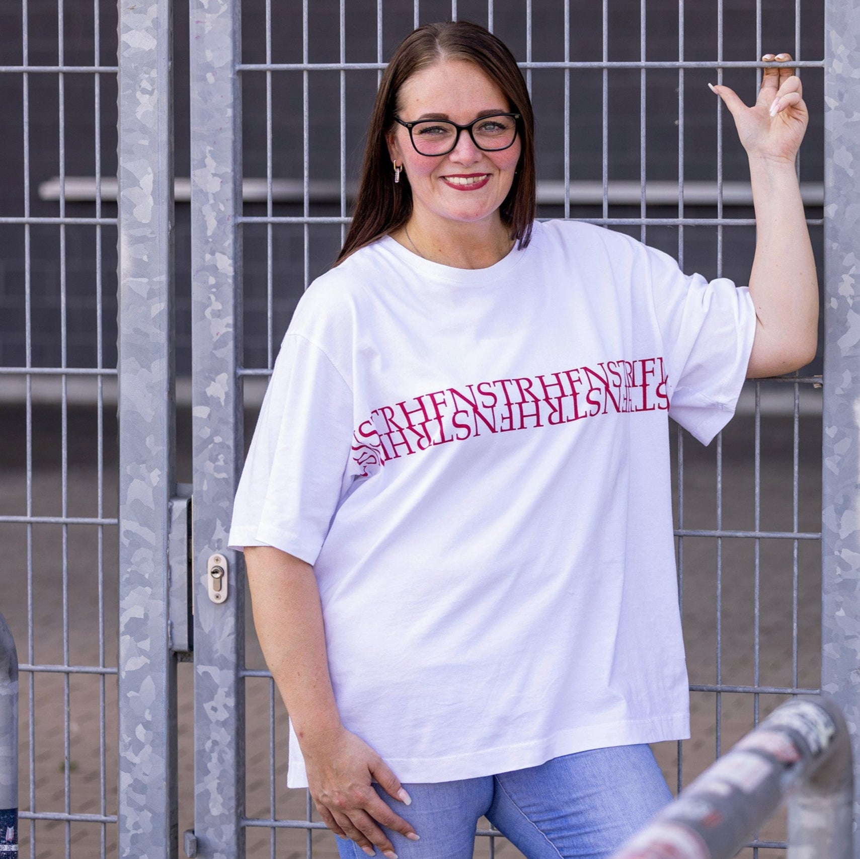 Jasmin trägt weißes Oversize Rundhals T-Shirt mit weißer Rippkante am Ausschnitt, rotes Nackenband, von Armloch zu Armloch roter Druck "HFNSTR"